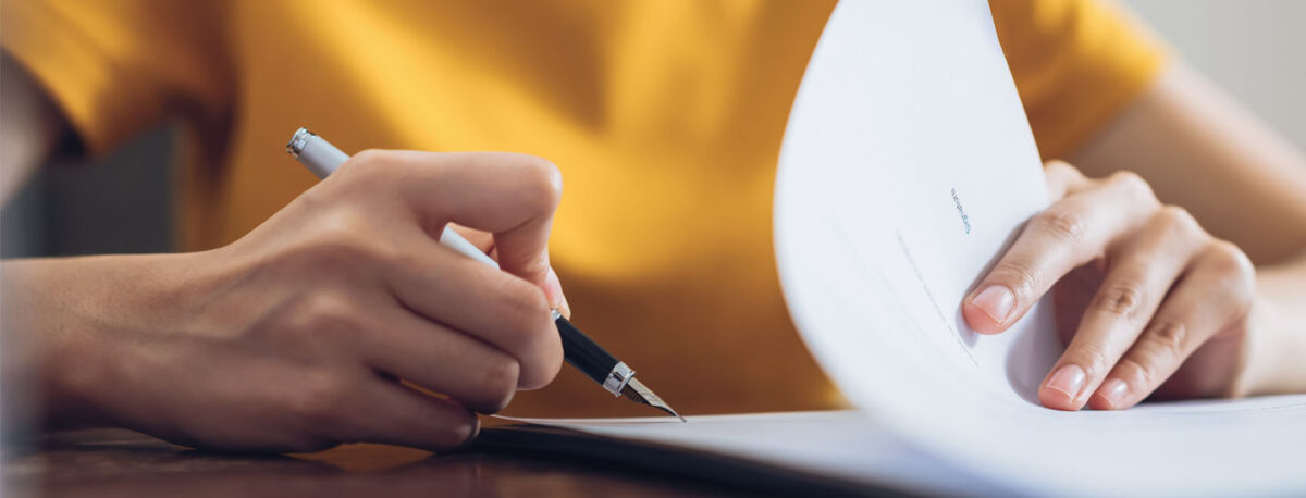A person in a yellow shirt writes on a document with a pen, holding and flipping through papers on a table.