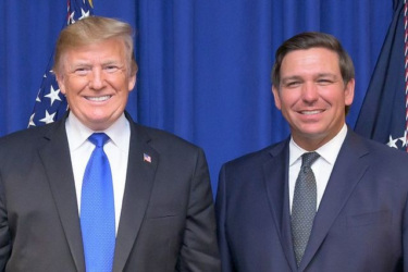 Two men in suits stand side by side, smiling, in front of blue curtains and American flags.