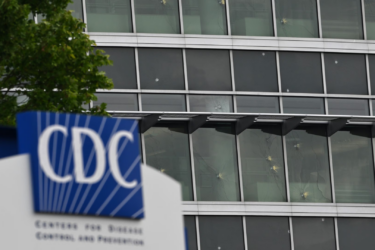 A sign with the CDC logo stands in front of a modern glass building with multiple windows; part of a tree is visible on the left side of the image.