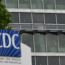 A sign with the CDC logo stands in front of a modern glass building with multiple windows; part of a tree is visible on the left side of the image.