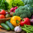 A colorful assortment of fresh vegetables, including carrots, cucumbers, tomatoes, bell peppers, broccoli, radishes, garlic, green beans, lettuce, kale, and spinach, arranged on a wooden surface outdoors.