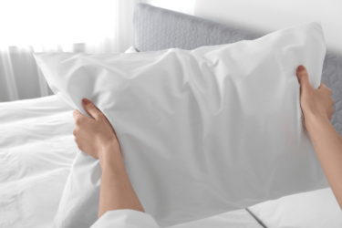 A person holds a white pillow with both hands in front of a neatly made bed with a gray headboard in a bright, sunlit bedroom.