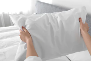 A person holds a white pillow with both hands in front of a neatly made bed with a gray headboard in a bright, sunlit bedroom.