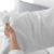 A person holds a white pillow with both hands in front of a neatly made bed with a gray headboard in a bright, sunlit bedroom.