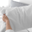 A person holds a white pillow with both hands in front of a neatly made bed with a gray headboard in a bright, sunlit bedroom.