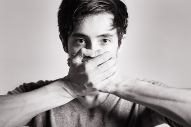 A young man looks directly at the camera, covering his mouth with both hands. The image is black and white, and the background is plain and blurred.