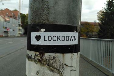 A sticker on a pole reads "I ♥ LOCKDOWN," with a heart symbol, on an empty street near buildings and trees.