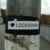 A sticker on a pole reads "I ♥ LOCKDOWN," with a heart symbol, on an empty street near buildings and trees.