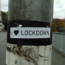 A sticker on a pole reads "I ♥ LOCKDOWN," with a heart symbol, on an empty street near buildings and trees.