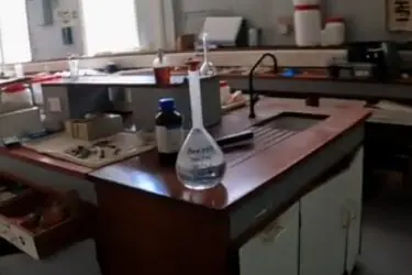 A laboratory with wooden workbenches, equipment, open drawers, bottles, and a labeled flask of clear liquid on the counter. The room appears well-lit with windows in the background.