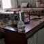 A laboratory with wooden workbenches, equipment, open drawers, bottles, and a labeled flask of clear liquid on the counter. The room appears well-lit with windows in the background.