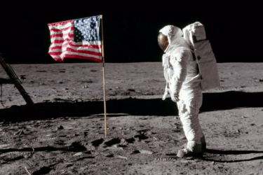 An astronaut in a white spacesuit stands on the Moon next to an American flag. The lunar surface is gray and rocky, with a black sky in the background. Boot prints are visible in the dust.