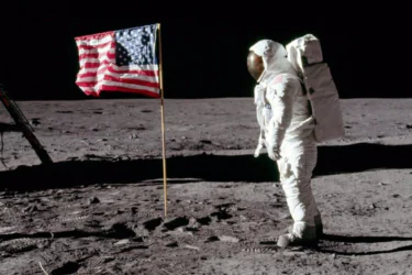 An astronaut in a white spacesuit stands on the Moon next to an American flag. The lunar surface is gray and rocky, with a black sky in the background. Boot prints are visible in the dust.