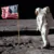 An astronaut in a white spacesuit stands on the Moon next to an American flag. The lunar surface is gray and rocky, with a black sky in the background. Boot prints are visible in the dust.