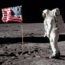 An astronaut in a white spacesuit stands on the Moon next to an American flag. The lunar surface is gray and rocky, with a black sky in the background. Boot prints are visible in the dust.