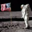 An astronaut in a white spacesuit stands on the Moon next to an American flag. The lunar surface is gray and rocky, with a black sky in the background. Boot prints are visible in the dust.