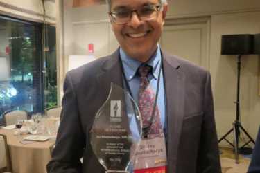 A man in a suit and patterned tie smiles while holding a glass award. He wears glasses and a conference badge that reads "Dr. Jay Bhattacharya" and "SPEAKER." The award is labeled "Frame of Freedom.