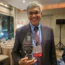 A man in a suit and patterned tie smiles while holding a glass award. He wears glasses and a conference badge that reads "Dr. Jay Bhattacharya" and "SPEAKER." The award is labeled "Frame of Freedom.