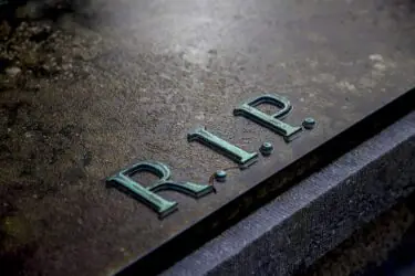 A close-up of a weathered gravestone with the letters "R.I.P." engraved on its surface, symbolizing rest in peace. The stone appears aged and has a slightly damp texture.