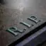 A close-up of a weathered gravestone with the letters "R.I.P." engraved on its surface, symbolizing rest in peace. The stone appears aged and has a slightly damp texture.