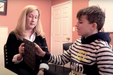 A woman holds a letterboard while a boy in a striped hoodie points at it with a pencil. They sit in a peach-colored room, engaged in a communication or learning activity.