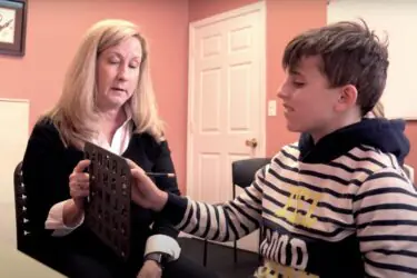 A woman holds a letterboard while a boy in a striped hoodie points at it with a pencil. They sit in a peach-colored room, engaged in a communication or learning activity.