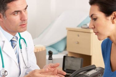 A doctor in a white coat talks with a woman in a blue top at a desk in a medical office, both appearing engaged in a serious conversation.