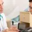 A doctor in a white coat talks with a woman in a blue top at a desk in a medical office, both appearing engaged in a serious conversation.
