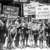 A group of boys holding protest signs advocating for justice and education, with messages like "We Want to Go to School" and "More School Less Hospital." The image appears to be from the early 20th century.
