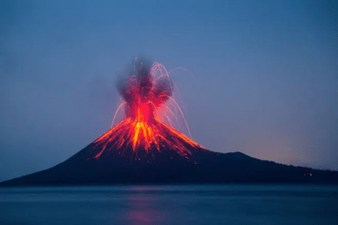 A volcano erupts at night, spewing bright orange lava and ash into the dark blue sky, with molten streams flowing down its slopes and a calm body of water in the foreground.