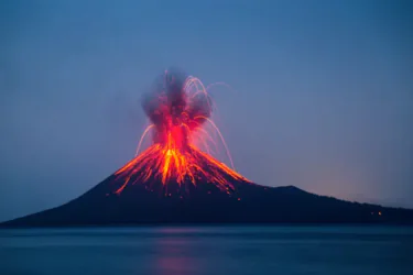 A volcano erupts at night, spewing bright orange lava and ash into the dark blue sky, with molten streams flowing down its slopes and a calm body of water in the foreground.