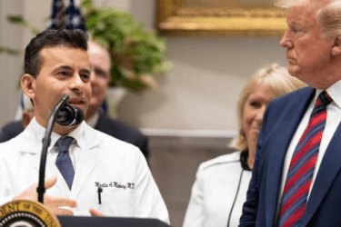 A doctor in a white coat speaks at a podium next to a man in a suit and striped tie, with others standing in the background.