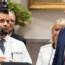 A doctor in a white coat speaks at a podium next to a man in a suit and striped tie, with others standing in the background.