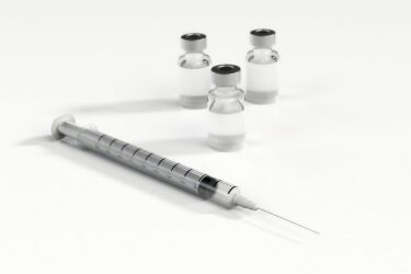 A syringe with a needle is placed in front of three small glass vials containing a clear liquid, all set on a white surface.