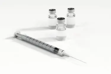 A syringe with a needle is placed in front of three small glass vials containing a clear liquid, all set on a white surface.