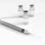 A syringe with a needle is placed in front of three small glass vials containing a clear liquid, all set on a white surface.