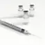 A syringe with a needle is placed in front of three small glass vials containing a clear liquid, all set on a white surface.