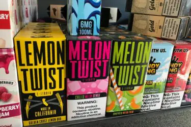 Colorful boxes of Lemon Twist and Melon Twist e-liquid are displayed on a store shelf. Warning labels and flavors are visible. Other vape products are partially visible on either side.