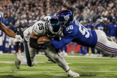 A football player in white is tackled by two players in blue uniforms during a game, with a crowd watching in the background.