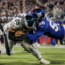 A football player in white is tackled by two players in blue uniforms during a game, with a crowd watching in the background.