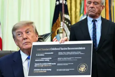A man in a suit holds a large chart with vaccine recommendations while sitting at a desk. Behind him stands another man in a suit. US flags and official emblems are visible in the background.