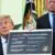 A man in a suit holds a large chart with vaccine recommendations while sitting at a desk. Behind him stands another man in a suit. US flags and official emblems are visible in the background.