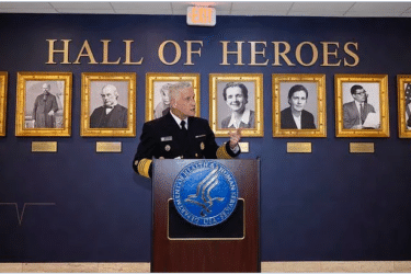 A uniformed official speaks at a podium in front of a “Hall of Heroes” wall, with framed portraits of notable individuals displayed behind him.