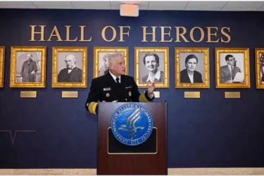 A uniformed official speaks at a podium in front of a “Hall of Heroes” wall, with framed portraits of notable individuals displayed behind him.