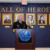 A uniformed official speaks at a podium in front of a “Hall of Heroes” wall, with framed portraits of notable individuals displayed behind him.