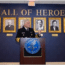 A uniformed official speaks at a podium in front of a “Hall of Heroes” wall, with framed portraits of notable individuals displayed behind him.