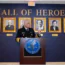 A uniformed official speaks at a podium in front of a “Hall of Heroes” wall, with framed portraits of notable individuals displayed behind him.