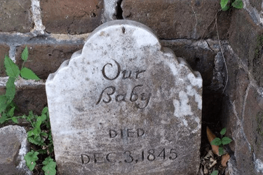 A weathered, white marble tombstone sits against a cracked brick wall. The inscription reads: “Our Baby” followed by “DIED DEC. 3, 1845.” Small green weeds and leaves grow in the dirt around the base of the headstone.