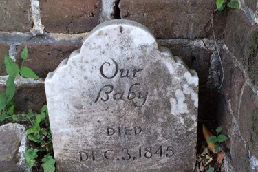 A weathered, white marble tombstone sits against a cracked brick wall. The inscription reads: “Our Baby” followed by “DIED DEC. 3, 1845.” Small green weeds and leaves grow in the dirt around the base of the headstone.