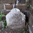 A weathered, white marble tombstone sits against a cracked brick wall. The inscription reads: “Our Baby” followed by “DIED DEC. 3, 1845.” Small green weeds and leaves grow in the dirt around the base of the headstone.